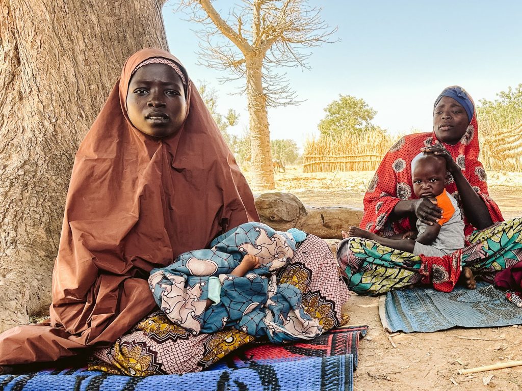 Two women hold young children in their laps while sitting on mats in Kano State, Nigeria.