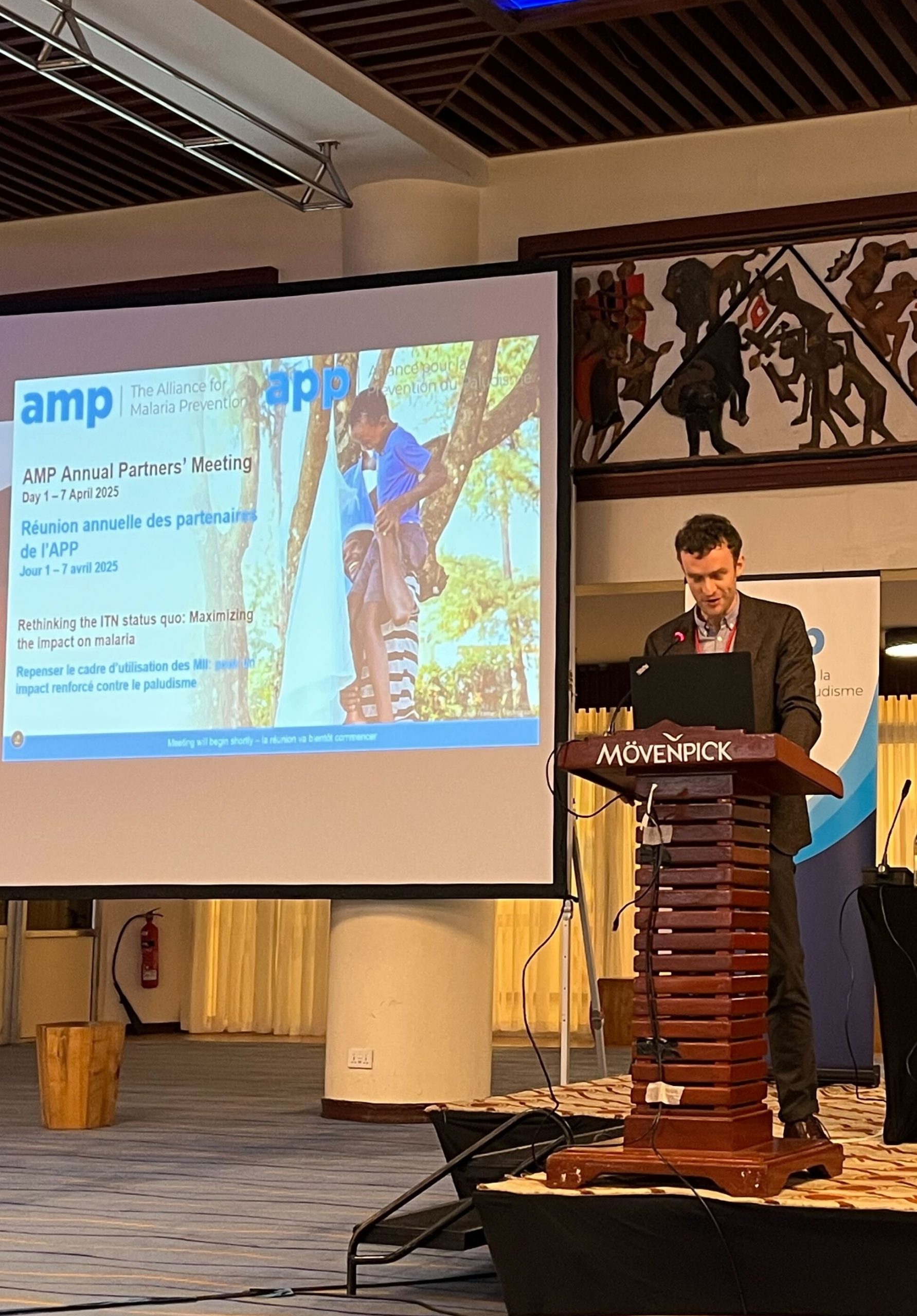 Alex Bowles speaks at the AMP Annual Partners’ Meeting beside a slide on improving the impact of insecticide-treated net programs.